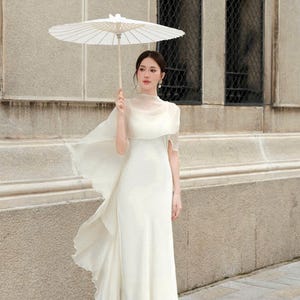 Könnte beinhalten: Ein weißes, bodenlanges Kleid mit einem transparenten, gerüschten Umhang und einem passenden Sonnenschirm. Das Kleid hat einen hohen Ausschnitt und einen fließenden Rock. Der Sonnenschirm ist weiß mit einem Holzgriff. Der Hintergrund ist ein Steingebäude.
