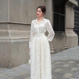 May include: A cream lace dress featuring a collared, button-down top and a long, flowing skirt. The dress has long sleeves and a fitted waist. The model is standing outside on a stone walkway.