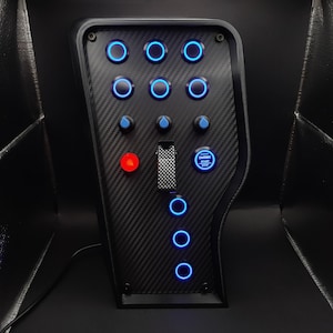 May include: A black carbon fiber control panel with multiple illuminated blue and red buttons, dials, and a toggle switch. The panel includes an "ENGINE START STOP" button.