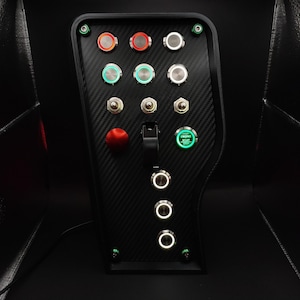 May include: A black carbon fiber textured control panel featuring a variety of metal push buttons, toggle switches, and a red emergency stop button. A central illuminated green engine start button is labeled with the text ENGINE START STOP.