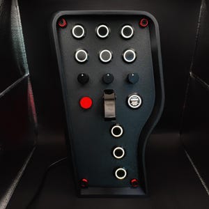 Peut inclure: Panneau de commande noir avec plusieurs boutons et interrupteurs. Comprend des anneaux blancs éclairés autour de certains boutons, un bouton rouge et un interrupteur à bascule. Le panneau porte l'inscription "ENGINE START STOP".