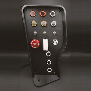 Puede incluir: Un panel negro con varios botones redondos y un botón rojo de parada de emergencia. Los botones están etiquetados con texto blanco y tienen luces LED blancas. El panel está montado en una superficie negra.