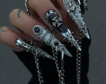 May include: Long, pointed stiletto-style artificial nails featuring a white and black color scheme. The nails are decorated with silver-toned metal spikes, small chains, cross emblems, and circular settings containing black stones, connected by silver chains.