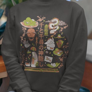 May include: A gray sweatshirt with a graphic print of characters and symbols from the Japanese animated film "Spirited Away". The text on the shirt reads "千と千尋の神隠し".