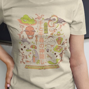 May include: Beige t-shirt featuring a colorful illustration with Japanese characters and various cartoon figures. The design includes a dragon, a frog, and other whimsical characters. The shirt is made of a soft material.