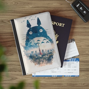 Op de afbeelding: Een witte paspoorthouder met een aquarel-illustratie van Totoro, een populair personage uit de Japanse animatiefilm Mijn buurman Totoro, die voor de skyline van Tokio staat. De illustratie toont een blauwe en grijze Totoro met een rode en witte Tokyo Tower op de achtergrond.