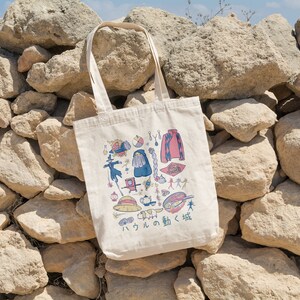 Peut inclure: Un sac fourre-tout en toile blanche avec une illustration color&eacute;e de divers personnages et objets du film d'animation japonais "Le Ch&acirc;teau ambulant". Le texte "Le Ch&acirc;teau ambulant" est &eacute;crit en japonais sur le sac.