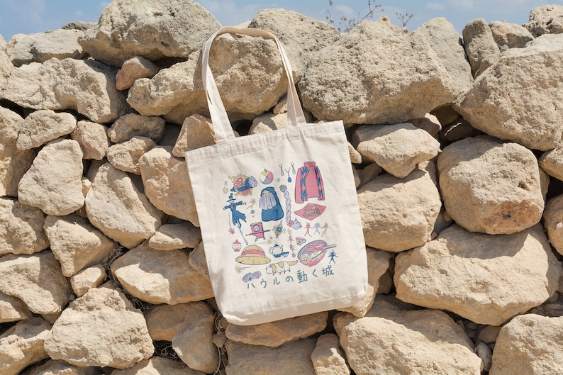 Peut inclure: Un sac fourre-tout en toile blanche avec une illustration color&eacute;e de divers personnages et objets du film d'animation japonais "Le Ch&acirc;teau ambulant". Le texte "Le Ch&acirc;teau ambulant" est &eacute;crit en japonais sur le sac.