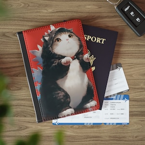 May include: A passport holder featuring a cartoon cat dressed in a Godzilla costume, set against a red background. The holder is next to a passport, plane tickets, and a car key. The passport has the word "SPORT" printed on it.