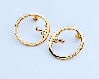May include: A pair of gold-toned circular earrings featuring a smooth, polished finish. Each earring consists of a thin, open hoop design with a decorative, fluid-like drip element extending from the inner edge toward the center of the circle.