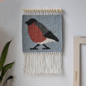 Puede incluir: Un tapiz tejido para colgar en la pared que representa un petirrojo estilizado con el pecho rojo, alas negras y detalles blancos. El fondo es de color azul claro y el tapiz tiene flecos blancos.