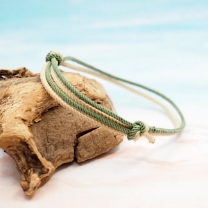 Op de afbeelding: Een handgemaakte armband met drie koorden in crème, olijfgroen en lichtgroen. De armband is aan de uiteinden geknoopt en rust op een stuk drijfhout. De achtergrond is zachtblauw en wit.