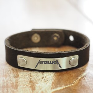 Op de afbeelding: Een zwart leren armband met een zilveren metalen plaat met daarop het woord "METALLICA" gegraveerd.