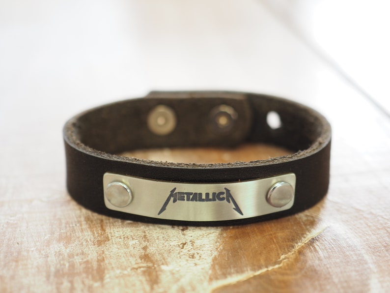Op de afbeelding: Een zwart leren armband met een zilveren metalen plaat met daarop het woord "METALLICA" gegraveerd.