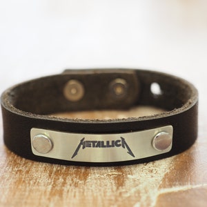 Op de afbeelding: Zwart leren armband met een zilveren metalen plaat gegraveerd met het woord "METALLICA" in een gestileerd lettertype.