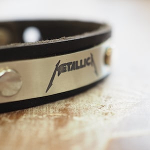 Op de afbeelding: Een zwart leren armband met een zilveren metalen plaat gegraveerd met het woord "METALLICA" in een gestileerd lettertype.