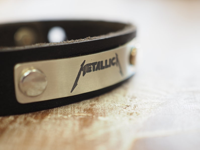 Op de afbeelding: Een zwart leren armband met een zilveren metalen plaat gegraveerd met het woord "METALLICA" in een gestileerd lettertype.