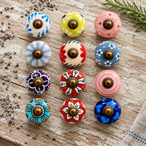 May include: A collection of twelve round ceramic cabinet knobs arranged in a grid. Each knob features a unique colorful pattern, including floral, geometric, and heart motifs, and is finished with a central brass-toned metal hardware piece.