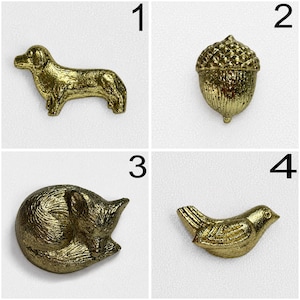 May include: A set of four gold-coloured metal decorative pins or buttons featuring distinct shapes: a standing dog, a textured acorn, a curled-up sleeping fox, and a small bird with wings tucked in, all displayed against a plain white background.