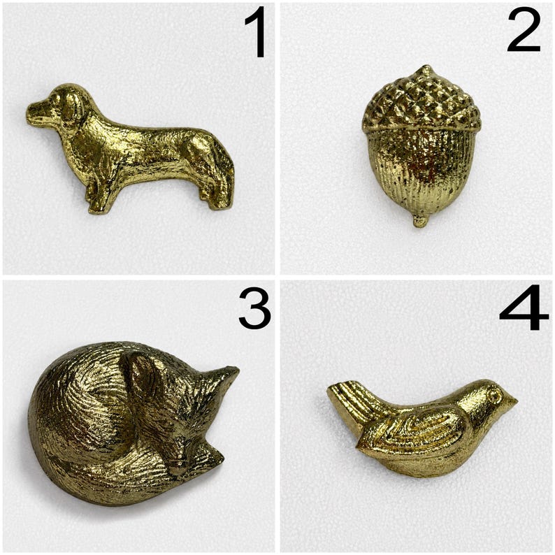 May include: A set of four gold-coloured metal decorative pins or buttons featuring distinct shapes: a standing dog, a textured acorn, a curled-up sleeping fox, and a small bird with wings tucked in, all displayed against a plain white background.