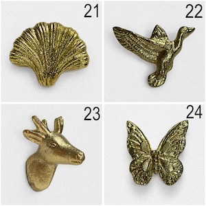 May include: A collection of four gold-toned metal decorative buttons or pins labelled 21 through 24. The items feature a ginkgo leaf, a flying bird, a deer head, and a butterfly, each with a textured, metallic finish and distinct natural shapes.