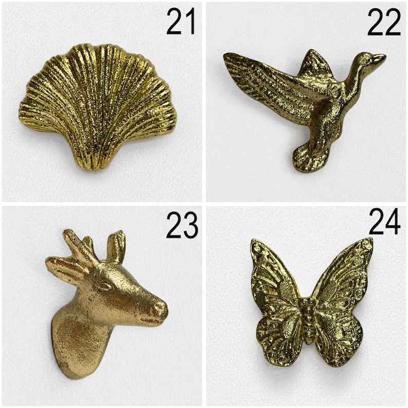 May include: A collection of four gold-toned metal decorative buttons or pins labelled 21 through 24. The items feature a ginkgo leaf, a flying bird, a deer head, and a butterfly, each with a textured, metallic finish and distinct natural shapes.
