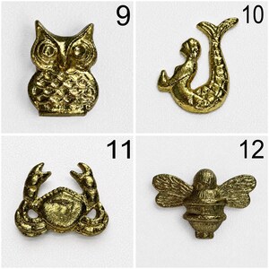 May include: A collection of four gold-toned metal charms labelled 9 through 12. The charms feature an owl, a mermaid, a crab, and a bee. Each piece has a textured, metallic finish and is displayed against a plain, light-coloured background.