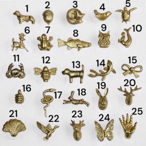 May include: A collection of twenty-five small gold-toned metal charms arranged in a grid, each numbered from 1 to 25. The charms feature various shapes including animals like a dog, owl, fish, and turtle, as well as objects like a bow, shell, and hand.