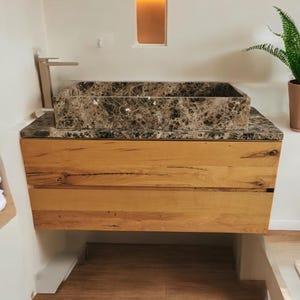 K&ouml;nnte beinhalten: Ein braunes Marmor-Waschbecken mit einem geb&uuml;rsteten Nickelhahn sitzt auf einem Holzwaschtisch mit zwei Schubladen. Der Waschtisch ist an einer wei&szlig;en Wand montiert.