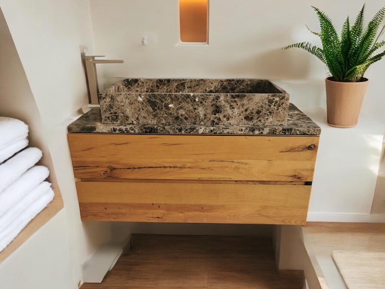K&ouml;nnte beinhalten: Ein braunes Marmor-Waschbecken mit einem geb&uuml;rsteten Nickelhahn sitzt auf einem Holzwaschtisch mit zwei Schubladen. Der Waschtisch ist an einer wei&szlig;en Wand montiert.