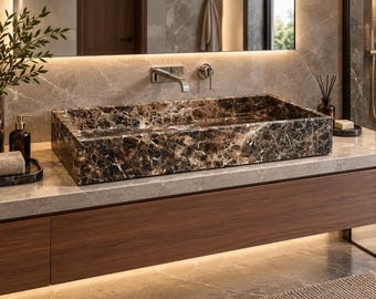 Puede incluir: Un lavabo rectangular de baño fabricado en mármol marrón oscuro con vetas blancas prominentes. La cubeta presenta un acabado pulido y está montada sobre una encimera de piedra gris claro sobre un mueble de baño de madera oscura.