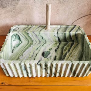 May include: A rectangular, green and white marble sink with a decorative, ridged edge. The sink has a circular drain and a tall, off-white faucet. The marble has a natural, flowing pattern.
