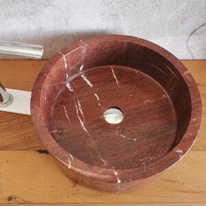 Op de afbeelding: Een ronde, rode marmeren wastafel met witte aderen. De wastafel heeft een gladde, gepolijste afwerking en een afvoergat in het midden.
