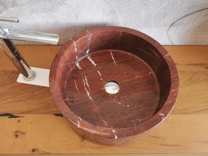 Op de afbeelding: Een ronde, rode marmeren wastafel met witte aderen. De wastafel heeft een gladde, gepolijste afwerking en een afvoergat in het midden.