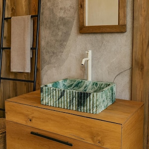 May include: A rectangular, green and white marble sink with a ribbed exterior sits atop a wooden cabinet. A white faucet is mounted above the sink. A wooden-framed mirror and a black towel rack are visible in the background.