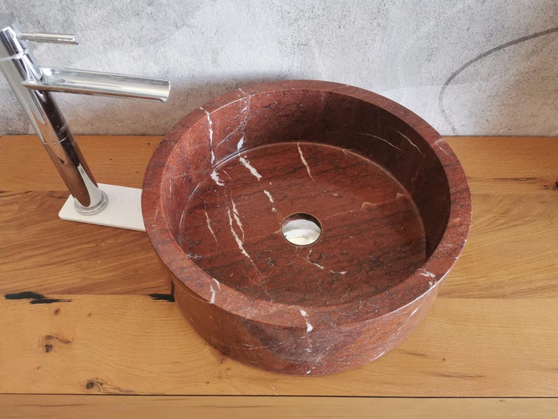 Op de afbeelding: Een ronde, rode marmeren wastafel met witte aderen. De wastafel heeft een gladde, gepolijste afwerking en een enkel afvoergat in het midden.
