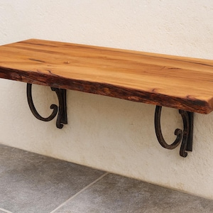 Könnte beinhalten: Ein schwebendes Holzregal mit natürlicher Kante, getragen von zwei verzierten, dunklen Metallhalterungen. Das Regal besteht aus hellbraunem Holz und ist an einer cremefarbenen Wand befestigt. Die Halterungen haben ein eingerolltes Design.