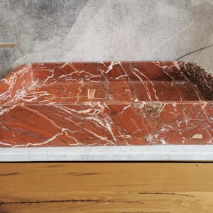May include: A rectangular red marble sink with white veining. The sink is made of natural stone and has a smooth, polished finish.