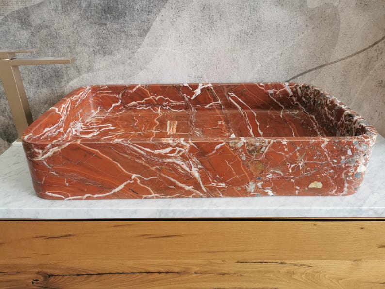 May include: A rectangular red marble sink with white veining. The sink is made of natural stone and has a smooth, polished finish.