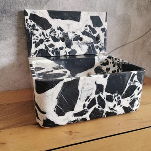 May include: A rectangular, open-top container with a hinged lid, crafted from black and white marble. The marble features a striking pattern of irregular shapes and veining. The container is set on a wooden surface.