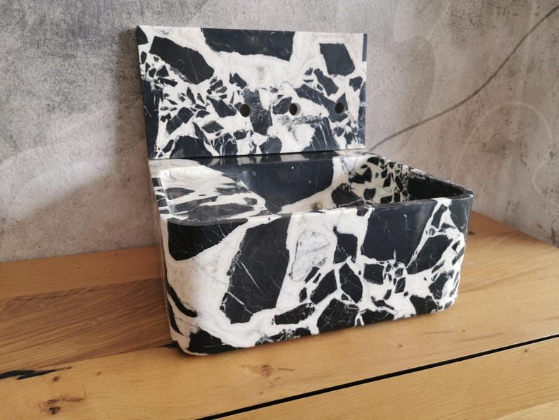 May include: A rectangular, open-top container with a hinged lid, crafted from black and white marble. The marble features a striking pattern of irregular shapes and veining. The container is set on a wooden surface.