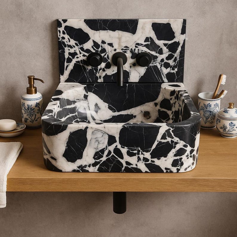 Un lavabo rettangolare in marmo bianco e nero con alzatina abbinata. Il rubinetto e le manopole sono nero opaco. Una mensola in legno sostiene il lavabo, sapone e accessori, tra cui un portaspazzolino e un piccolo barattolo con motivi floreali blu. il lavabo è realizzato in marmo grand antique, monoblocco, scavato da un blocco di marmo