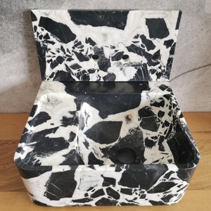 May include: A black and white marble-patterned soap dish with a hinged lid. The dish has a rectangular base with rounded corners and a divided interior. The lid features a similar pattern and has several small holes.