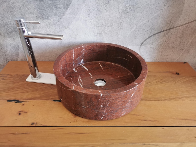Op de afbeelding: Een ronde, rode marmeren wastafel met witte aderen. De wastafel staat op een houten aanrecht.