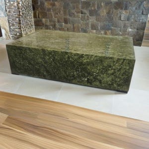 May include: A large, rectangular coffee table made of green stone with a rough, textured surface. The table is sitting on a light beige tiled floor with a wooden floor in the foreground.