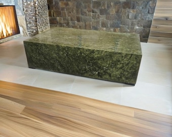 Puede incluir: Una mesa de centro grande y rectangular hecha de piedra verde con una superficie rugosa y texturizada. La mesa está sobre un suelo de baldosas beige claro con un suelo de madera en primer plano.