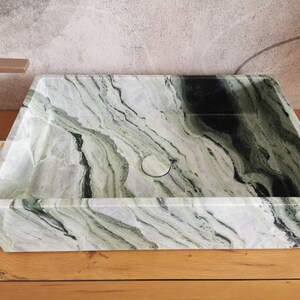 May include: Rectangular marble sink with a natural green and white veined pattern. The sink has a centered drain and is set on a wooden surface. The faucet is a brushed gold color.