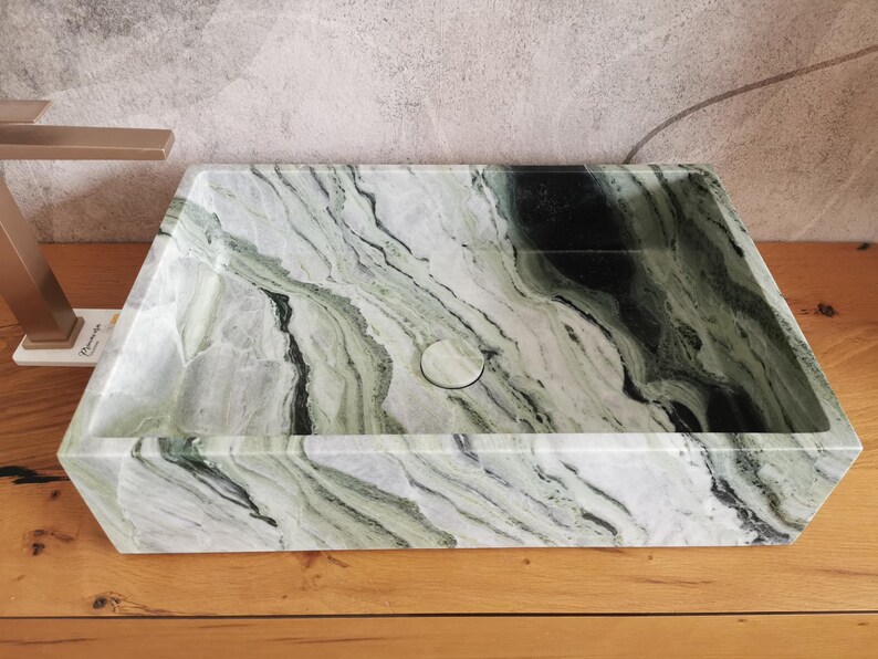 May include: Rectangular marble sink with a natural green and white veined pattern. The sink has a centered drain and is set on a wooden surface. The faucet is a brushed gold color.