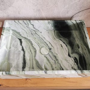 May include: Rectangular marble sink with a natural green and white veined pattern. The sink has a central drain and is set on a wooden surface. The design features a smooth, polished finish.