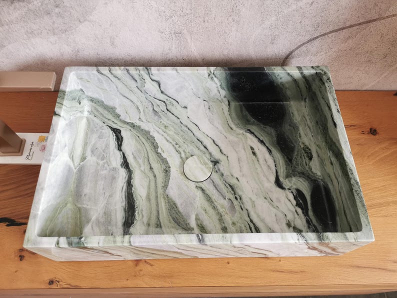 May include: Rectangular marble sink with a natural green and white veined pattern. The sink has a central drain and is set on a wooden surface. The design features a smooth, polished finish.